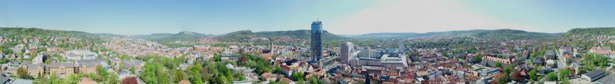 Panoramabild des Stadtkerns Jena, aufgenommen aus der Höhe des JenTowers. Im Zentrum der JenTower, FSU Campus und die Stadtmitte mit Kirche und Marktplatz. An den Rändern Wohngebiete.