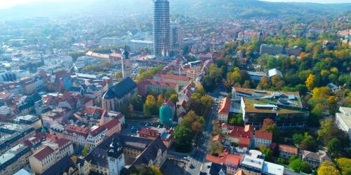 Schräges Luftbild mit Blick auf das Jenaer Zentrum. Zu sehen sind der JenTower, die Neue Mitte, eine große universitäre Bibliothek, die historische Stadtkirche Sankt Michael und das Universitätshauptgebäude.