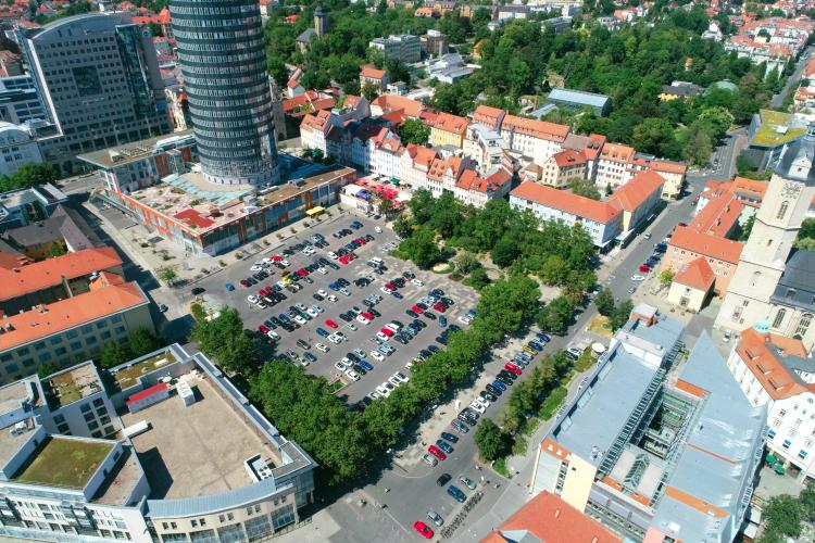 Autoparkplatz der in U-Form von Bäumen umrandet wird. Daneben das kleine Einkaufszentrum Neue Mitte.