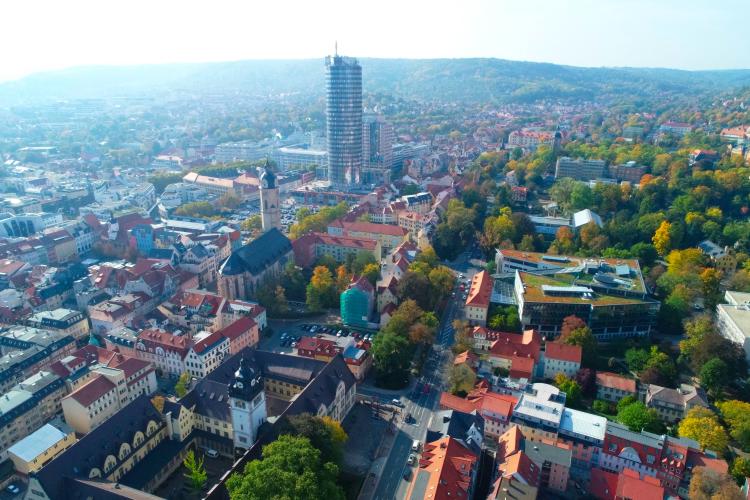 Schräges Luftbild mit Blick auf das Jenaer Zentrum. Zu sehen sind der JenTower, die Neue Mitte, eine große universitäre Bibliothek, die historische Stadtkirche Sankt Michael und das Universitätshauptgebäude.