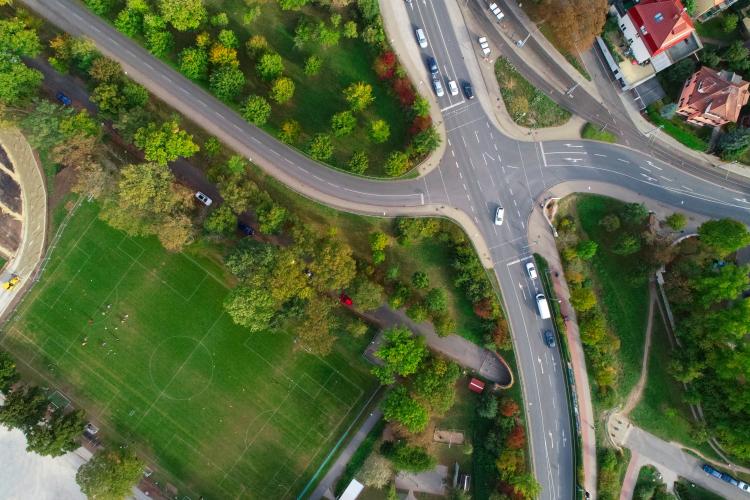 Eine große Straßenkreuzung, auf der ein paar Autos fahren. Daneben hinter einem schützenden Baumwall ein Fußballfeld, wo gerade eine Mannschaft trainiert.