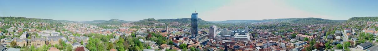 Panoramabild des Stadtkerns Jena, aufgenommen aus der Höhe des JenTowers. Im Zentrum der JenTower, FSU Campus und die Stadtmitte mit Kirche und Marktplatz. An den Rändern Wohngebiete.